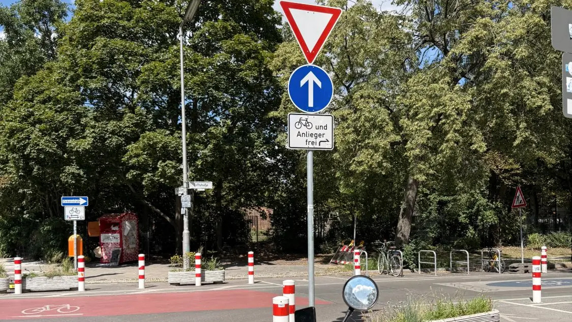 A sign stack at a Berlin junction: a give way triangle, a one way arrow, and a plate letting bicycles and residents through, with a red cycle lane crossing in front.